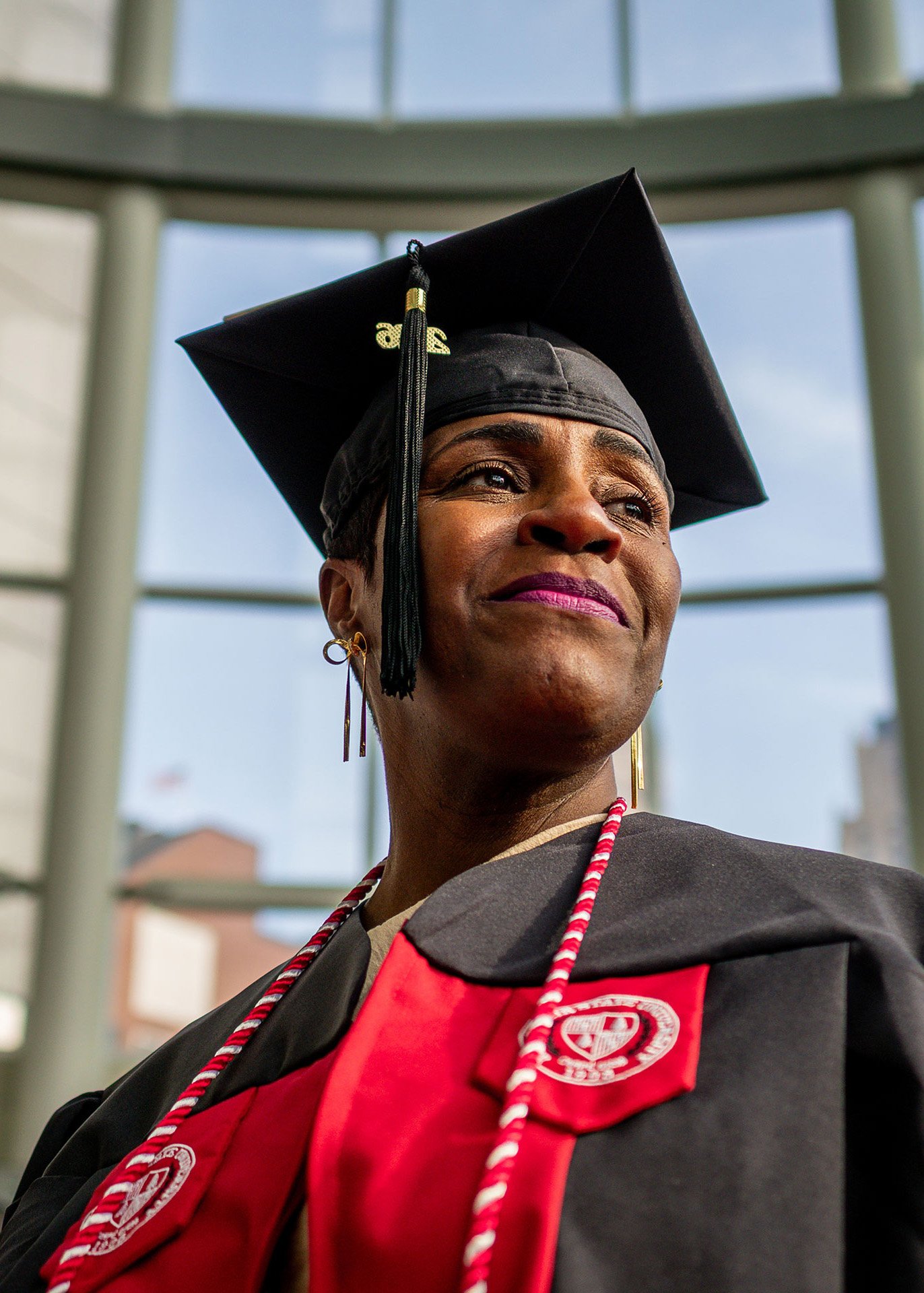 A Montclair State University graduate wearing a black cap and gown with red honors cords looks off into the distance with a proud expression in the bright glass atrium of Prudential Center during the 2026 Spring Commencement ceremony.