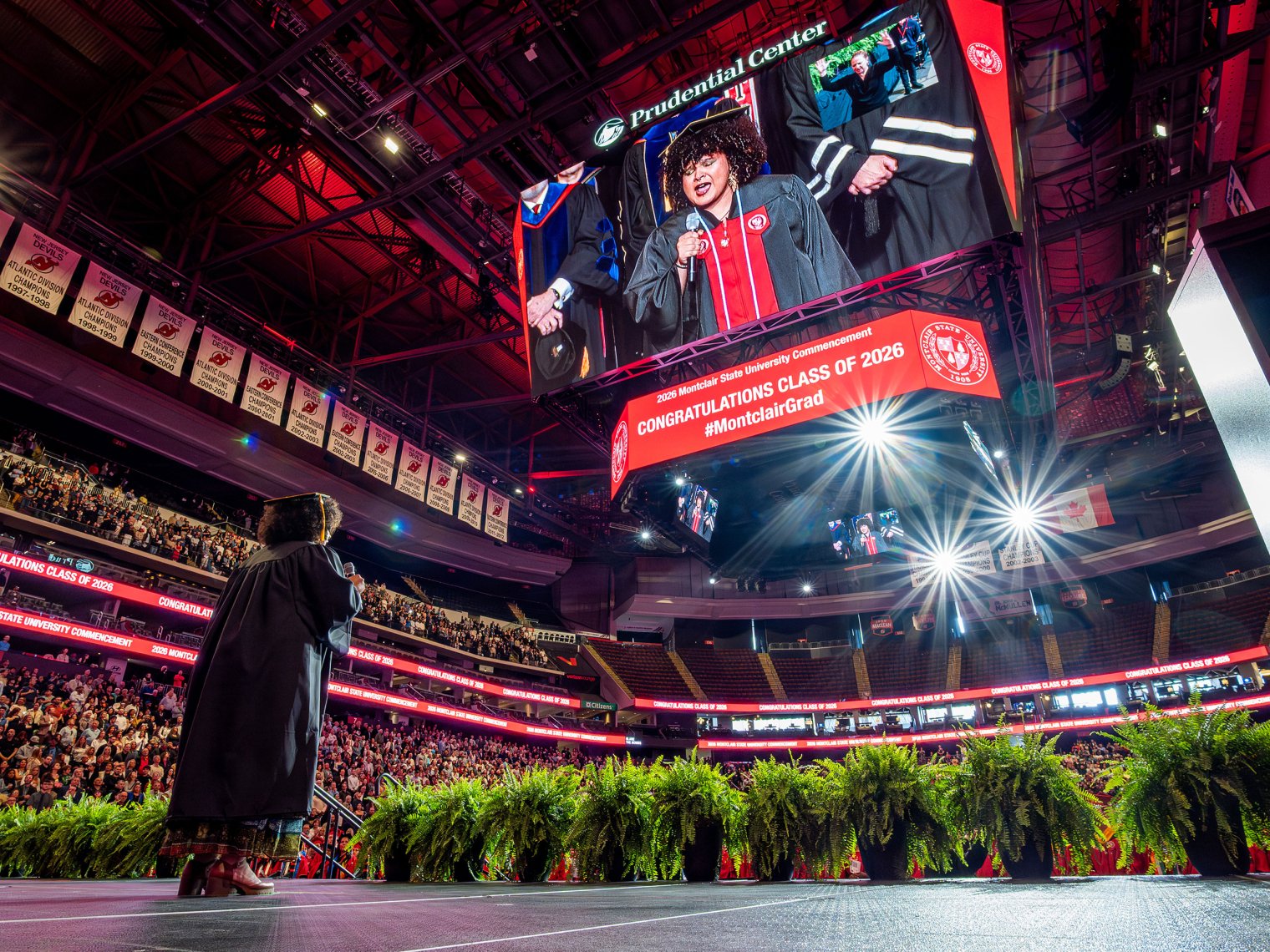 Montclair State University student Arianna Amina Joseph stands on the arena stage singing the national anthem into a microphone as the audience rises, while her image appears on the large Prudential Center video board above a banner reading “Congratulations Class of 2026.”