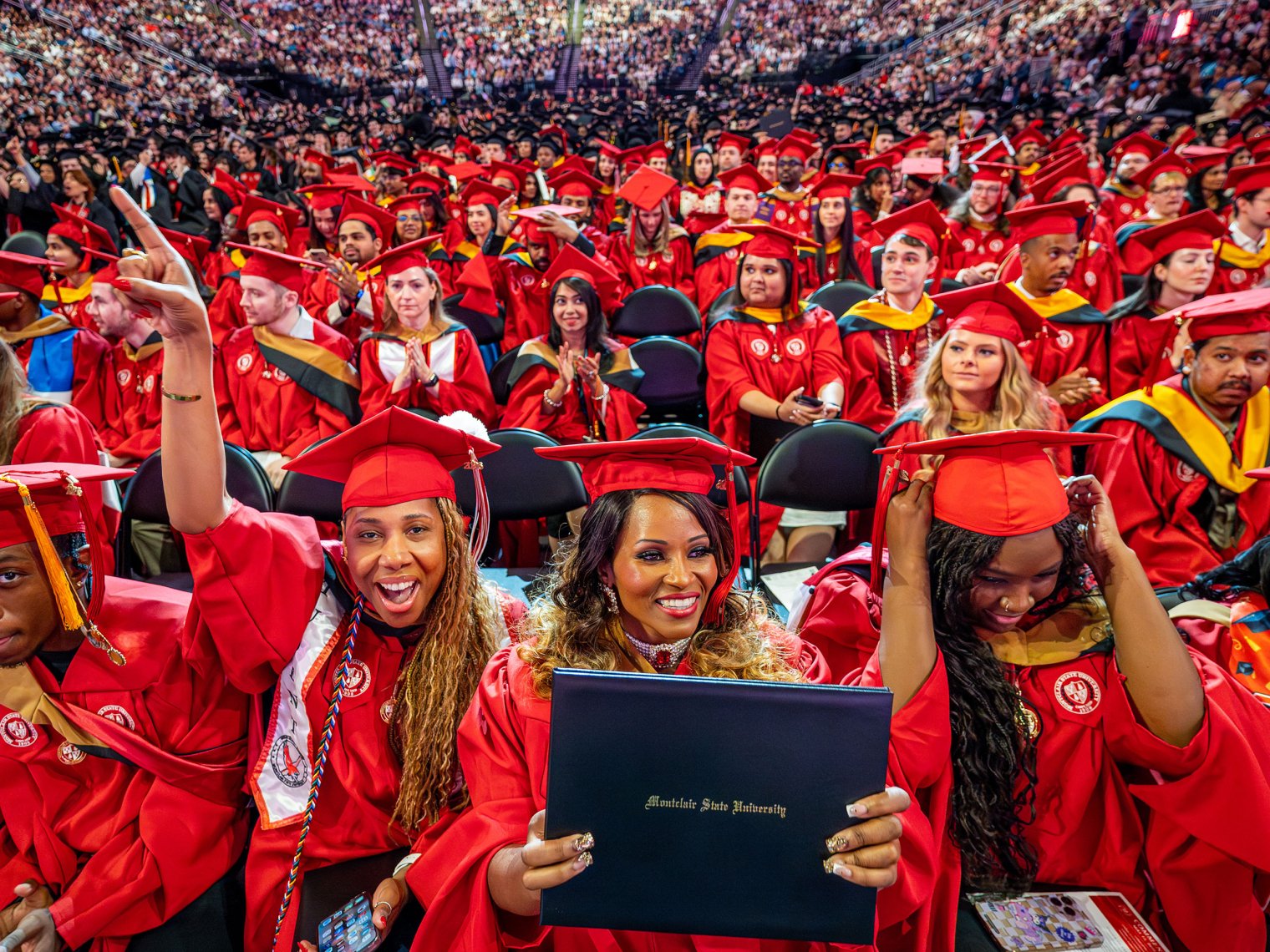A large group of Montclair State University Graduate School students in bright red caps and gowns smile, cheer and hold up diplomas while seated together on the arena floor during the 2026 Spring Commencement ceremony, with rows of fellow graduates and guests filling the stands behind them.