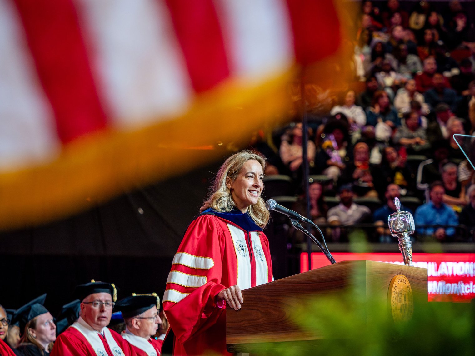 New Jersey Governor Mikie Sherrill, wearing red and white academic regalia, speaks at a podium during Montclair State University’s 2026 Spring Commencement, with a large American flag blurred in the foreground.