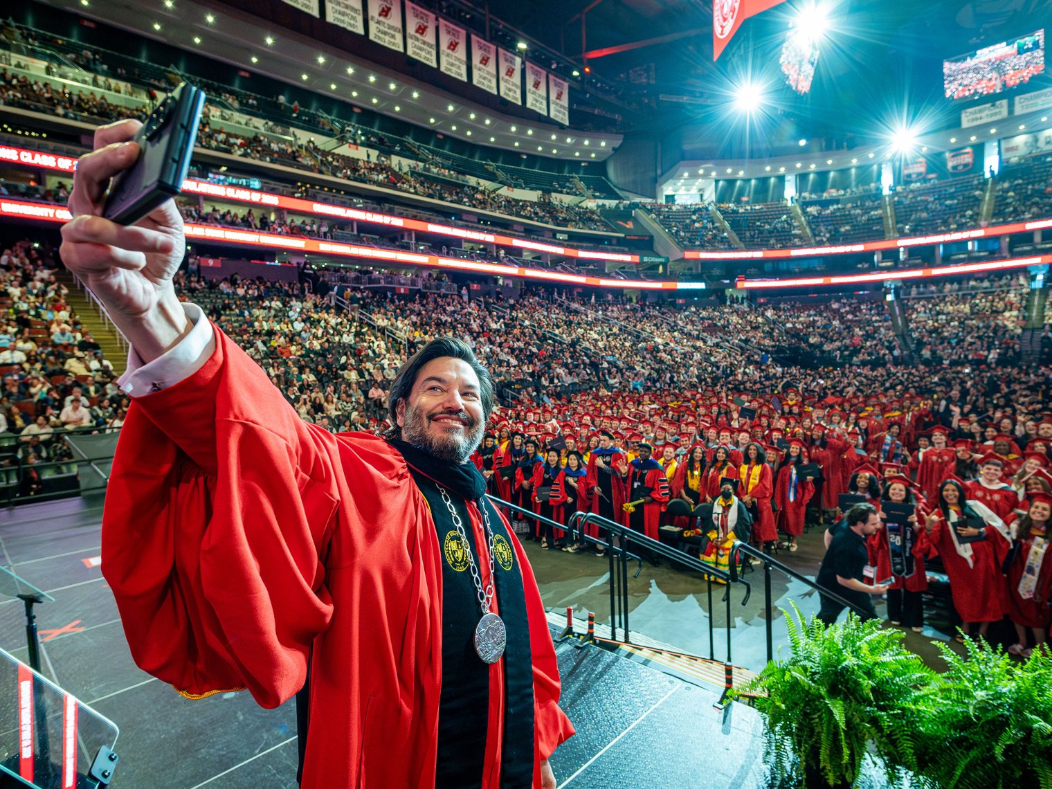 President Jonathan Koppell in red academic regalia holds up a phone to take a selfie with rows of graduating students in red gowns and a full arena of guests behind them during Montclair State University’s 2026 Spring Commencement.