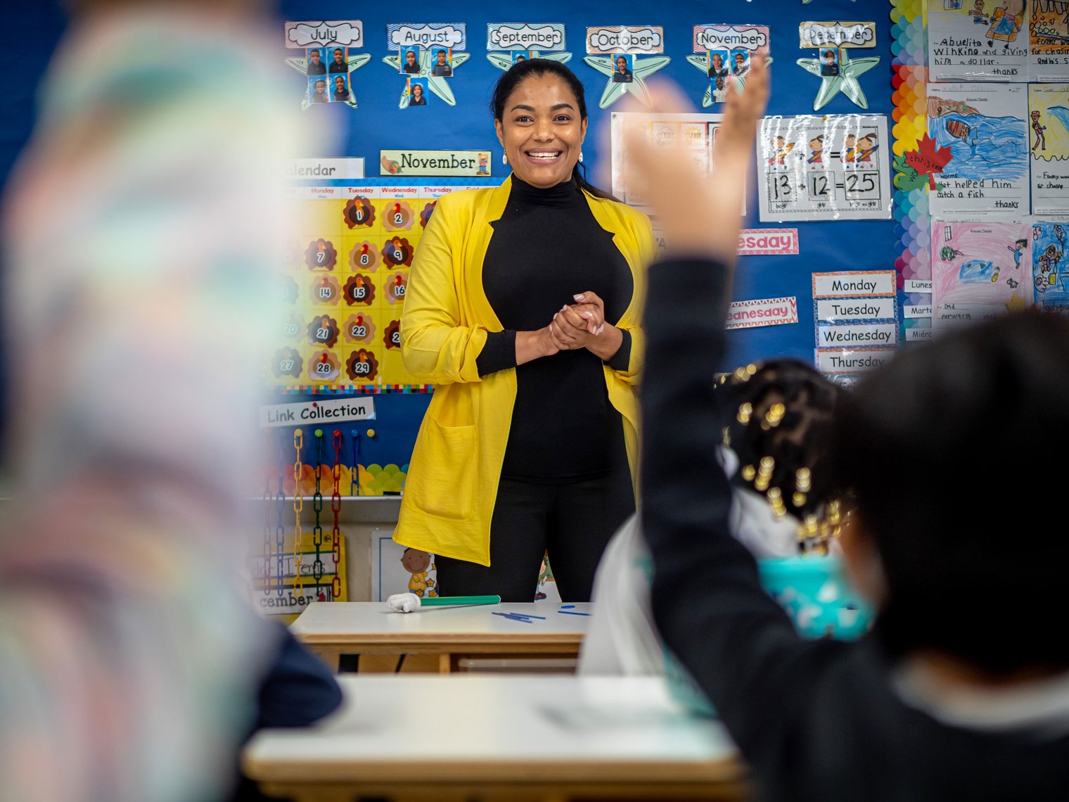 Teacher in yellow blazer at front of classroom facing young students