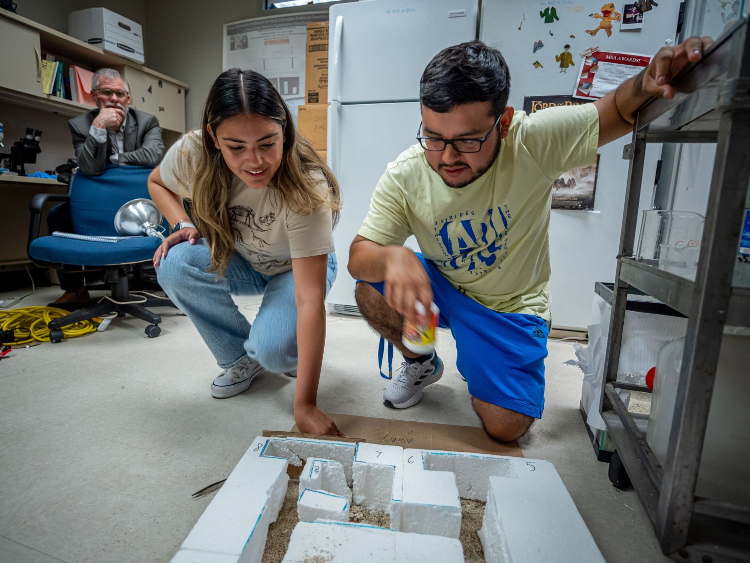 Two students kneel behind a small square maze. Their professor sits behind them.