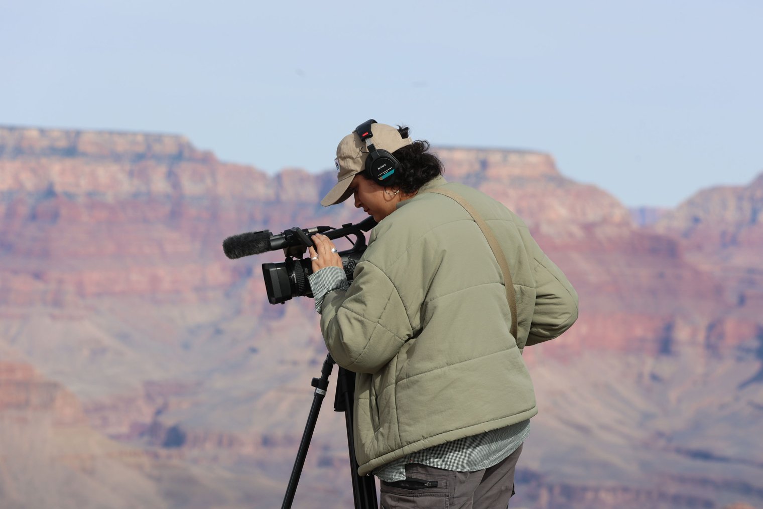 Dani Mazariegos stands outdoors on rocky terrain, wearing a light jacket and headset, as she adjusts a video camera on a tripod with desert cliffs and canyon walls in the background.