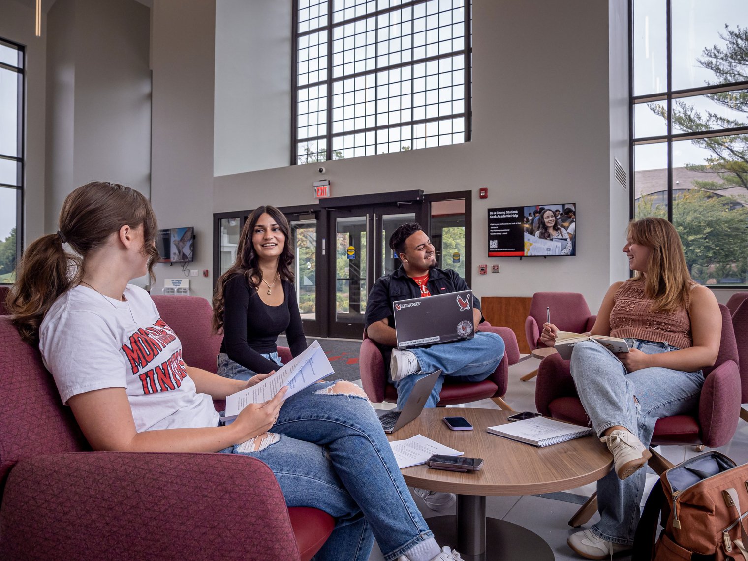 Four students sitting on couches. They are talking amongst themselves as they write notes and one is using their laptop.
