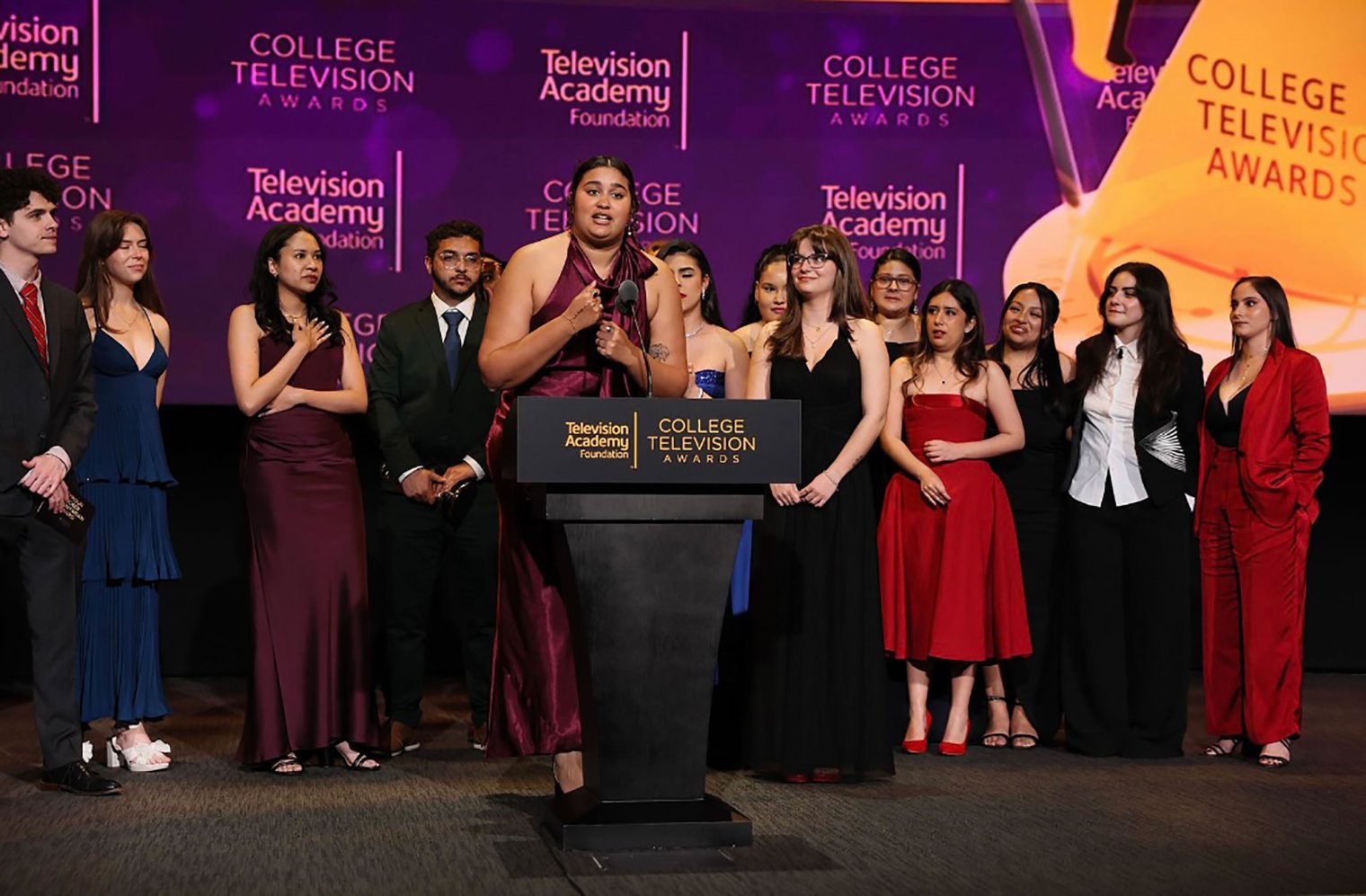Onstage at the College Television Awards, Dani Mazariegos stands at a podium labeled “Television Academy Foundation College Television Awards,” speaking into a microphone while holding an award, surrounded by Montclair co-reporters dressed in formal evening wear against a purple awards‑show backdrop.