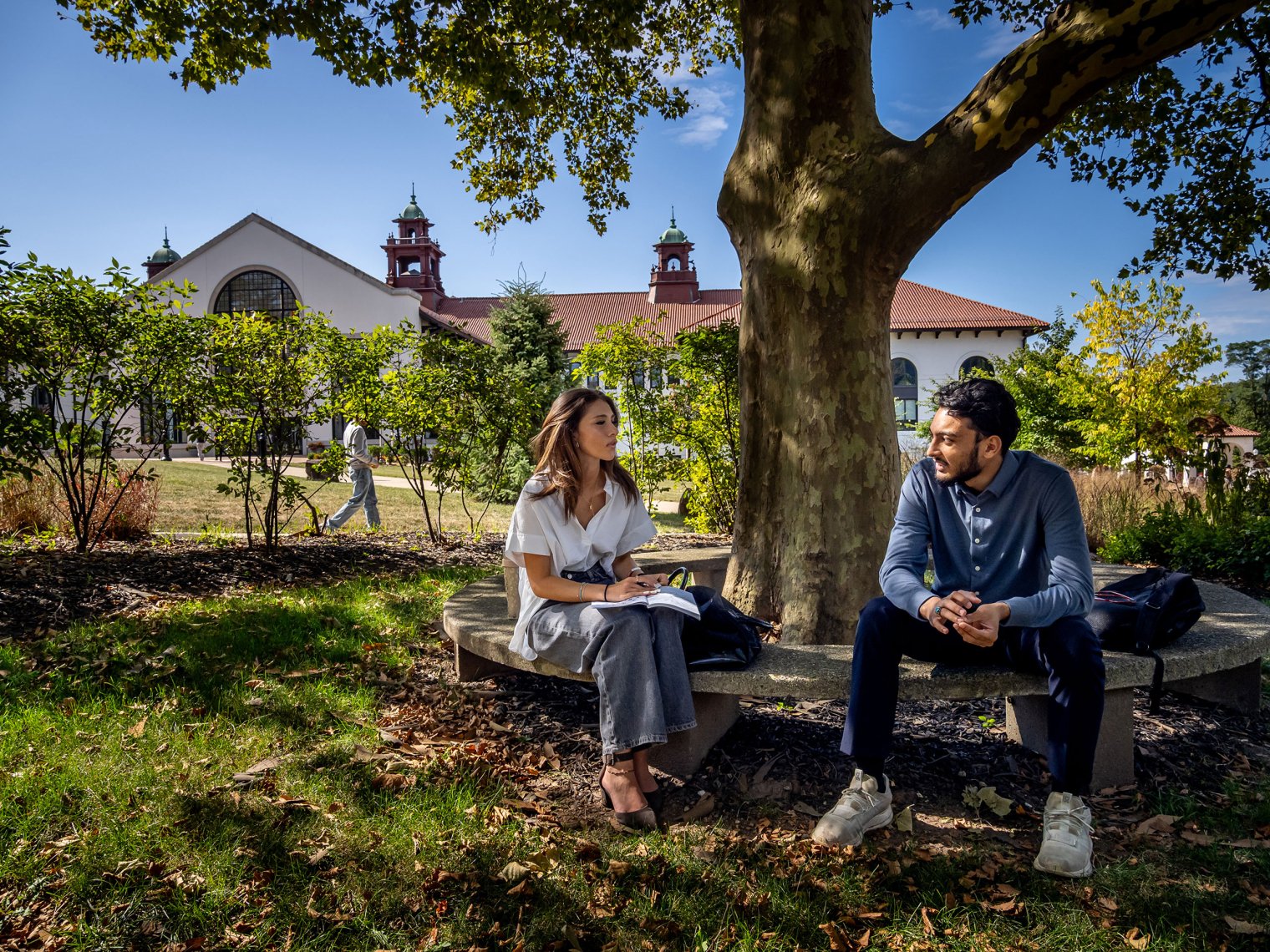 Two students talking outside on a bench that wraps around a tree.