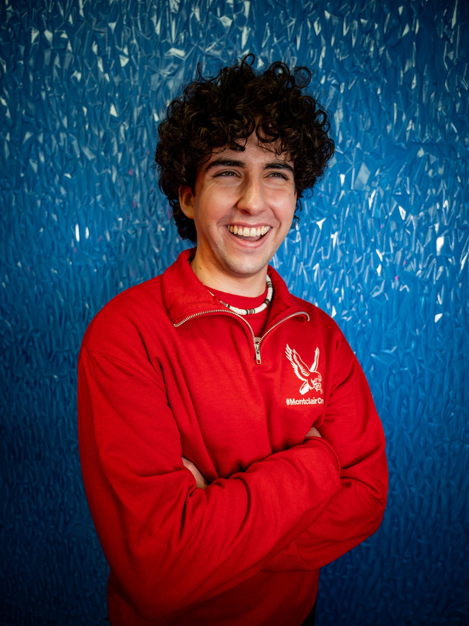 Photo of a man with his arms crossed and a goofy grin while wearing a bright red Montclair Athletics sweater