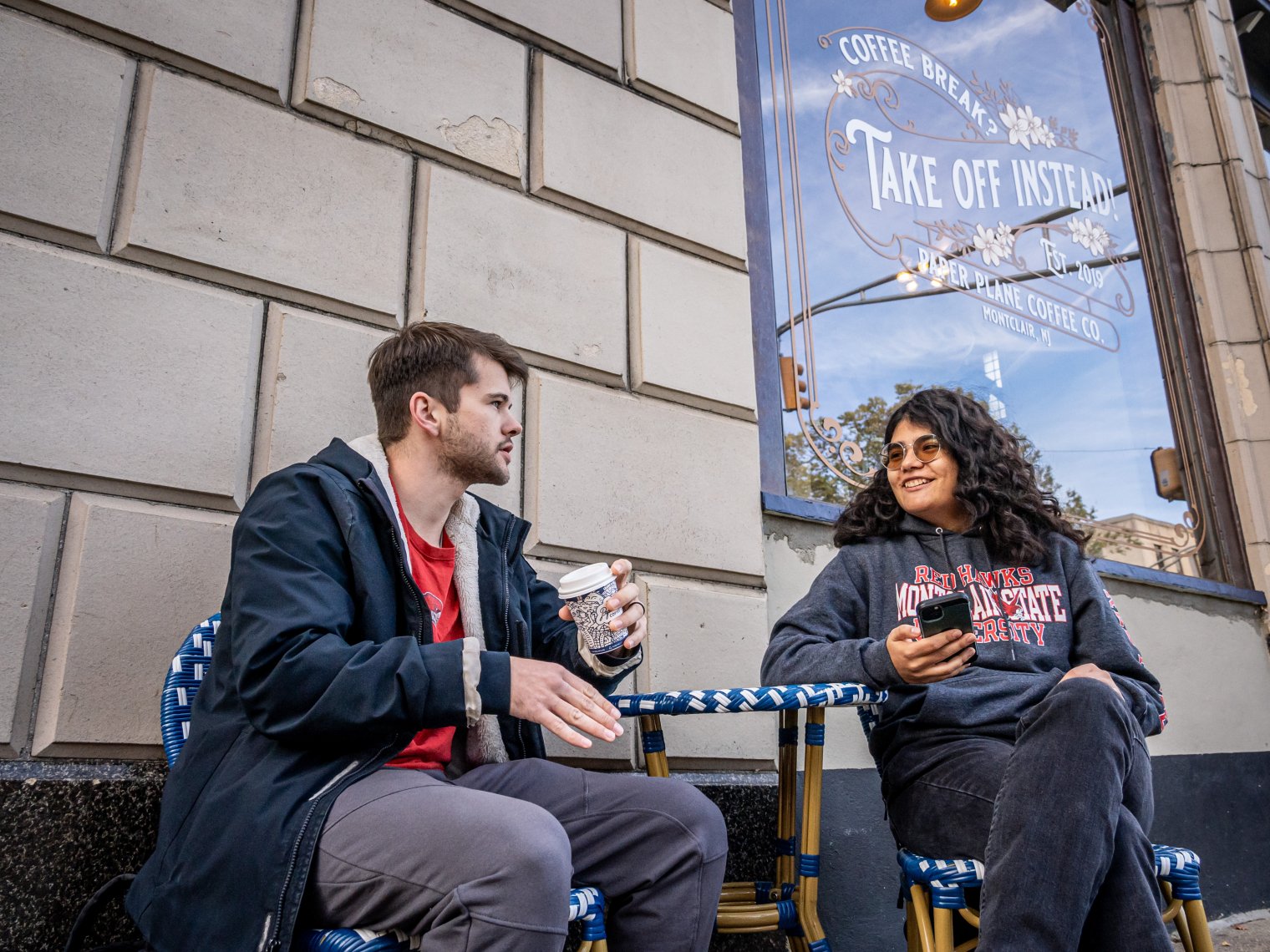 Two students chat while sitting at a coffee shop’s outdoor table. 