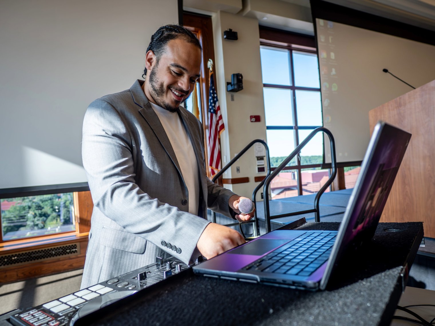 Ashon Lanada, wearing a gray blazer, sets up a laptop and sound equipment on stage, with a screen behind him displaying Montclair State University and partner college logos.