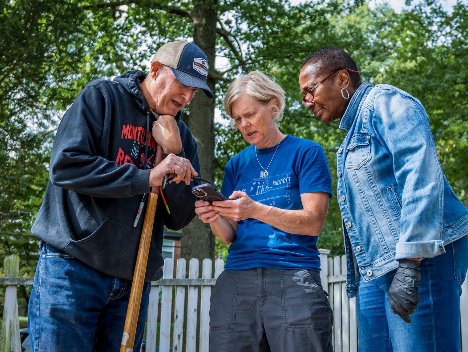 A man leaning on a shovel and two women confer while looking at a cell phone.