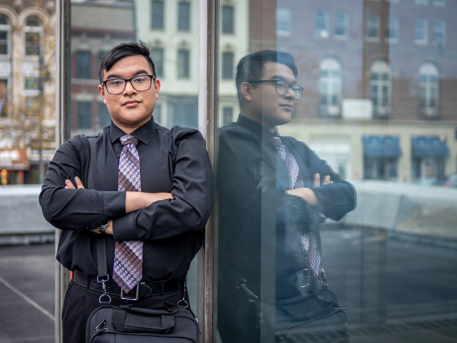 A student poses against an office building, with his arms cross, reflected in a glass wall.
