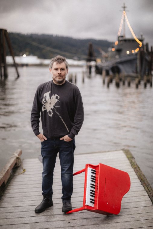 Gabriel Kahane stands on a pier with a small red piano tipped on its side near his feet