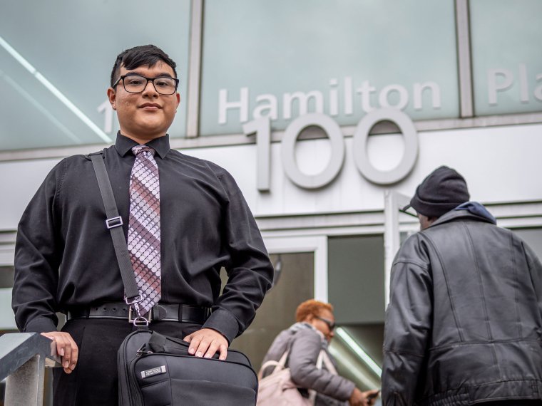 Montclair student Jose Carlos Aguilar wearing a black shirt and patterned tie posing in front of an office building