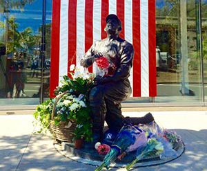 Photo of Yogi Berra statue in front of Yogi Museum with flowers at its base and an american flag in the background